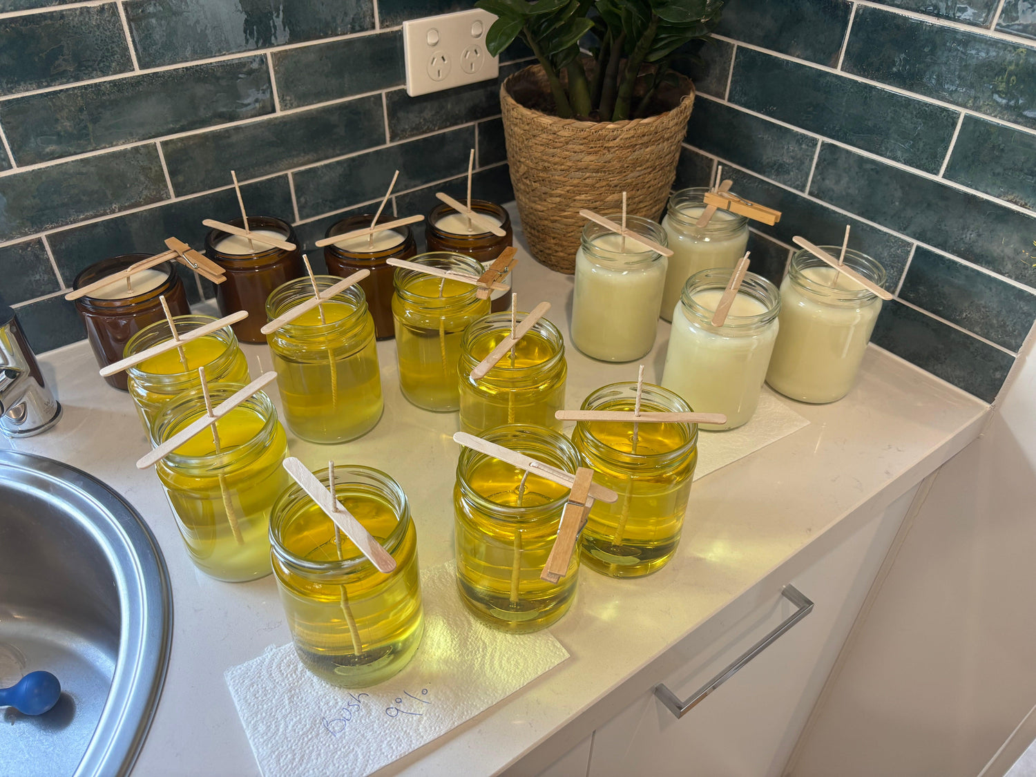 Hand-poured soy candles in glass jars with wooden wicks, set on a kitchen counter with green tile backsplash.