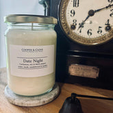Date Night soy candle in glass jar with silver lid, featuring cardamom, iris & black pepper notes, beside vintage clock